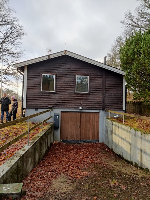 Äldre mörkbrunt fritidshus med garage under, löv på marken, visar slitage, personer i bakgrunden.