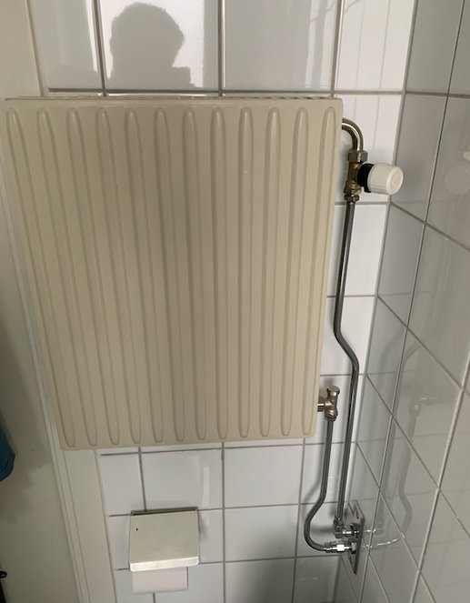 Vit vägghängd radiator i ett trångt badrum med vita kakelväggar och termostatventil.