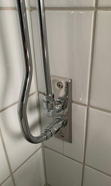 Närbild av en handdukstork monterad på en kaklad badrumsvägg med synliga vattenrör och anslutningar.