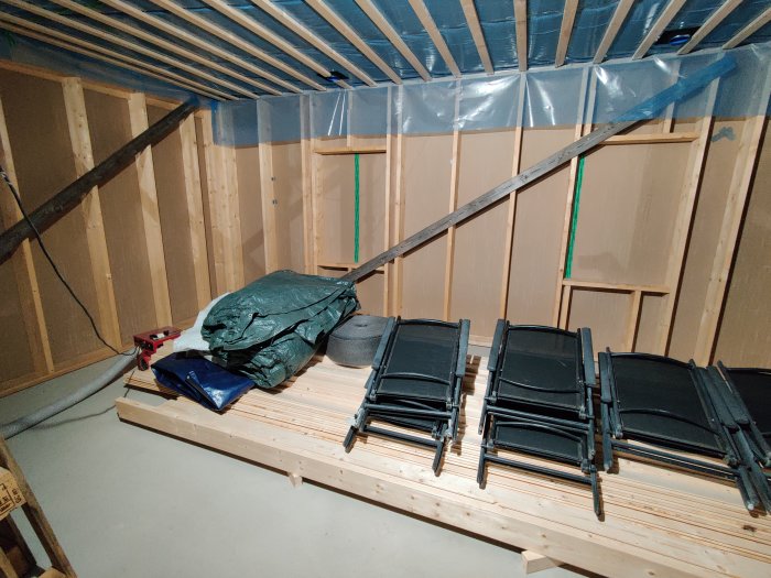 Underkonstruktion av ett rum med synliga träbjälkar och plast, staplar av byggmaterial och avfuktare.