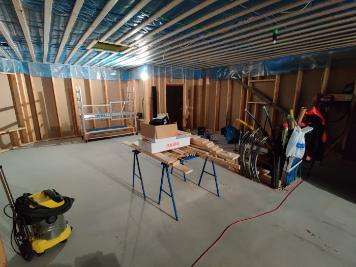 En ouppvärmd källare under renovering med byggmaterial, verktyg och en dammsugare på golvet.