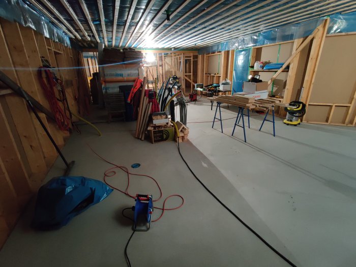 Byggplats inomhus med isolering under tak, byggmaterial och verktyg spridda över betonggolv.