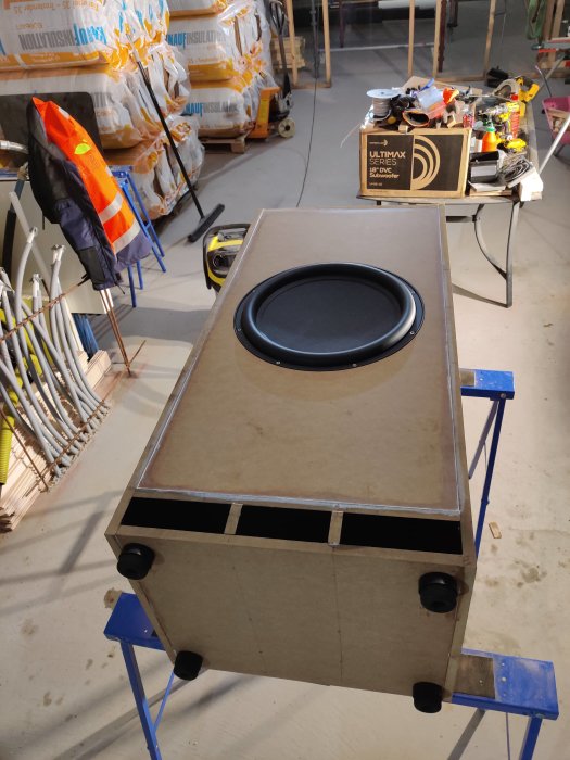 Stor, nybyggd högtalarkabinett i plywood monterad på arbetsbockar i ett rörigt byggrum.