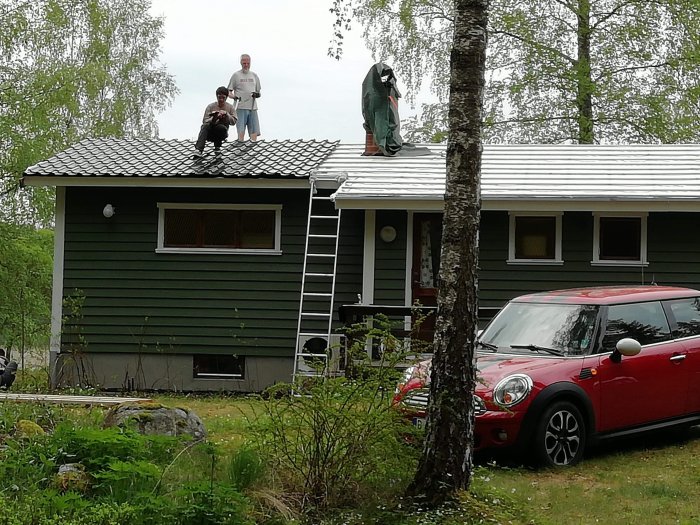 Personer på taket av ett grönt fritidshus arbetar med takrenovering, med en röd bil parkerad framför huset.