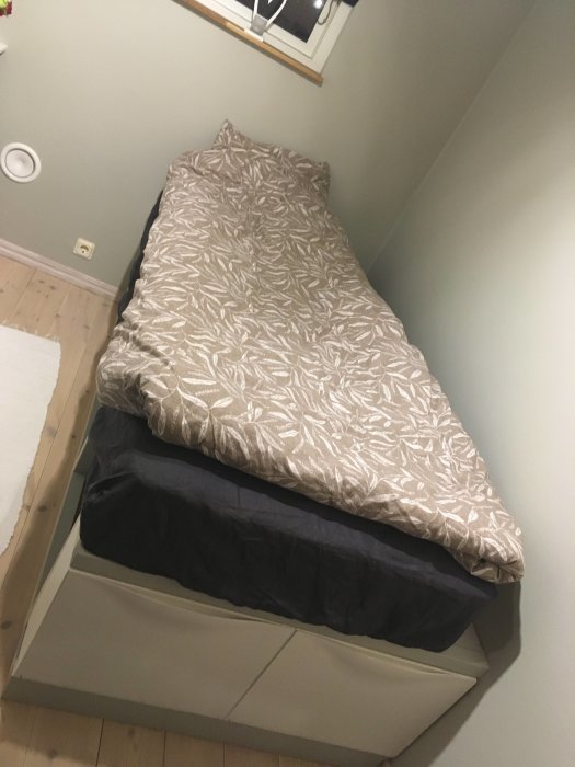 Hemmagjord säng med träram och integrerade förvaringslådor vid fotänden, täckt av ett mönstrat överkast.