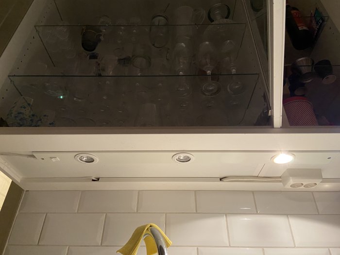 Spotlights monterade under överskåp med vit kakelvägg och en glashylla med glasögon i bakgrunden.