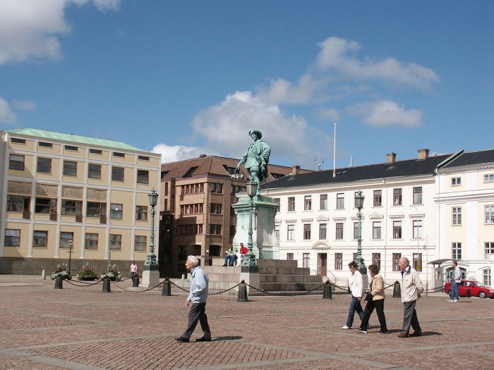 Stadstorg med människor som promenerar, staty i centrum och äldre byggnader i bakgrunden.