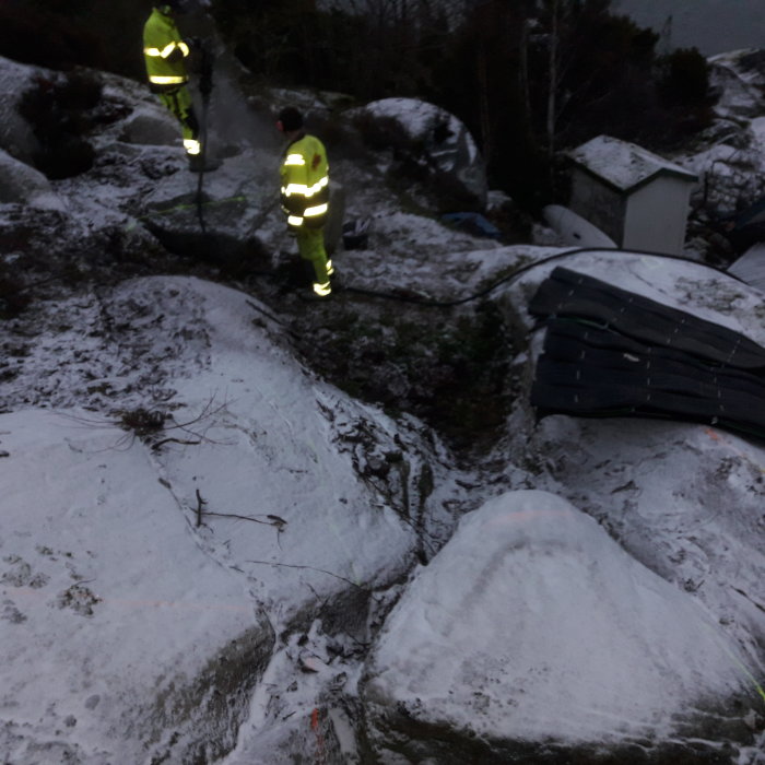 Två personer i reflexkläder på ett snöbeklätt byggområde under skymning.