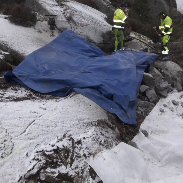 Arbetspersonal i säkerhetskläder täcker sten och rasmassor med en blå presenning utomhus.