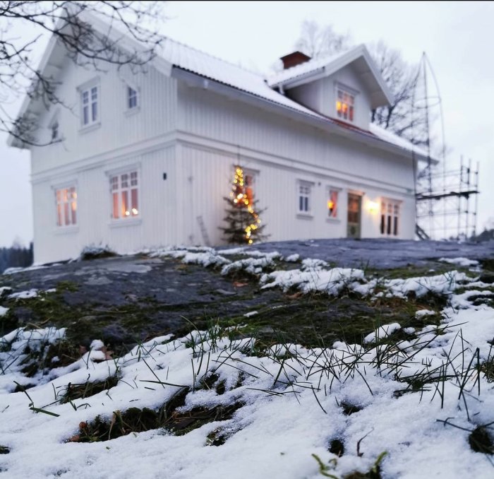 Vit villa med julbelysning och lätt snötäcke, framför gräs och snö på marken.