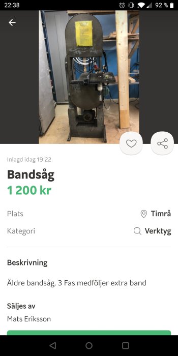 Äldre bandsåg på golvet i ett verkstadsrum med synliga rör och verktyg i bakgrunden.