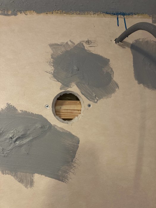 Spacklad vägg med ett runt hål visande träreglar bakom, intill en böjd plastslang och några kladdiga märken.