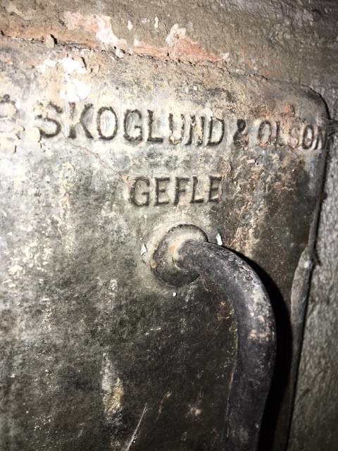 Närbild på en fastrostad lucka i skorsten med texten "SKOGLUND & OLSON GEFLE" och ett olöst handtag.