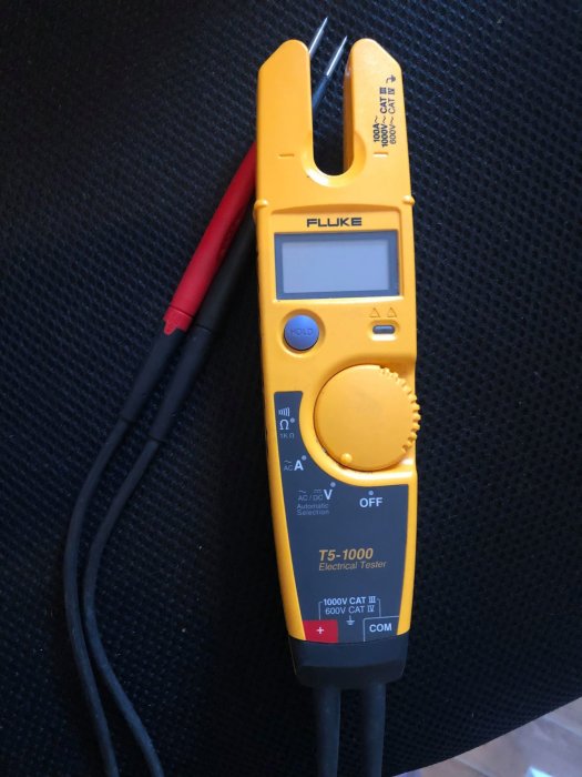 Fluke T5-1000 elektrisk testare med klämmor och digital display på svart bakgrund.