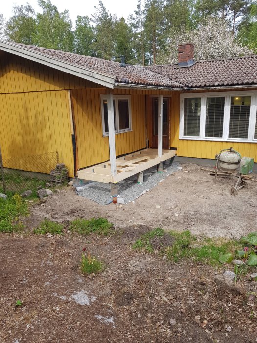 Påbörjad utbyggnad av gult hus med ny grund och träram under konstruktion, med presenningar och grusig mark runtomkring.