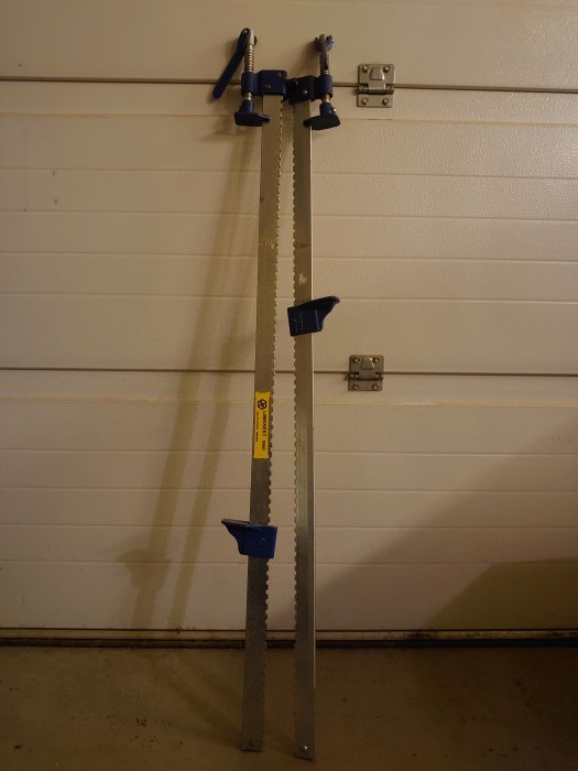 Två begagnade stångsågar med blå handtag lutade mot en vit garagevägg, inköpta för ett DIY-projekt.