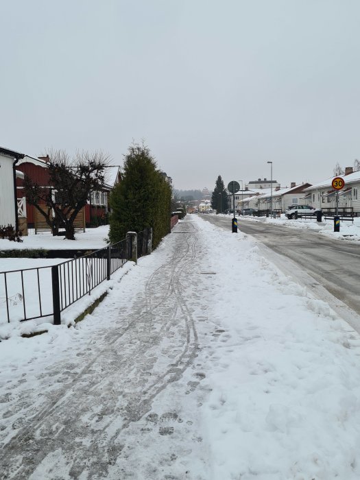 Snötäckt gata i bostadsområde med fotspår och hus i bakgrunden, hastighetsskylt 30 km/h synlig.