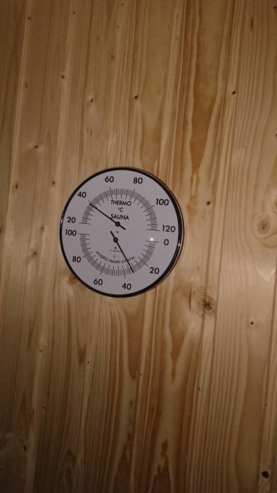 Termometer och hygrometer på trävägg i bastu som visar temperatur och luftfuktighet.