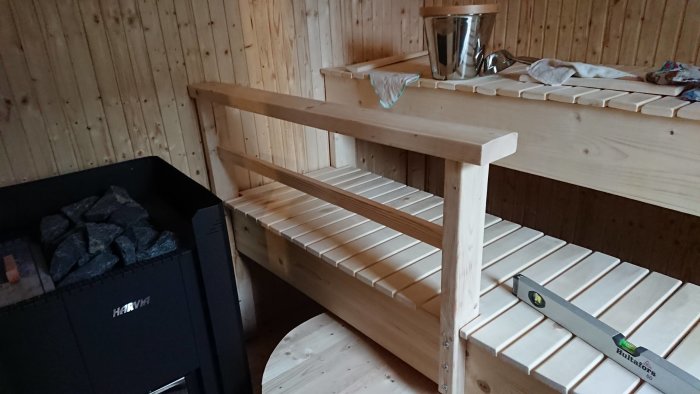 Inre delen av en bastu med träbänkar och en Harvia Linear 22 LS kamin med stenar, närbild på en vattenbalja med termometer.