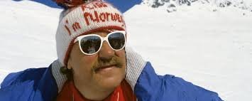 Person med skidmössa och solglasögon ler mot kameran framför snötäckt bergslandskap.