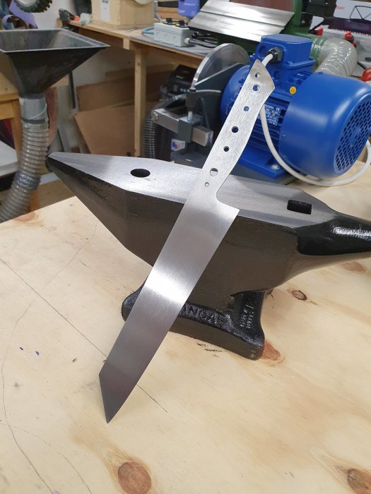 Ett knivblad lutar mot ett litet 20kg ambolt på ett träbord i en verkstad.