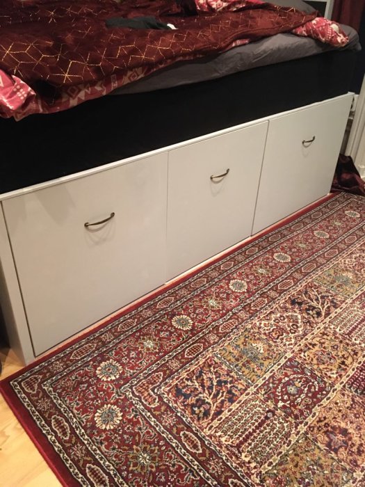 Hemmabyggd förvaring under säng, med två vita trådbackar och en stor låda, på ett orientaliskt mattgolv.