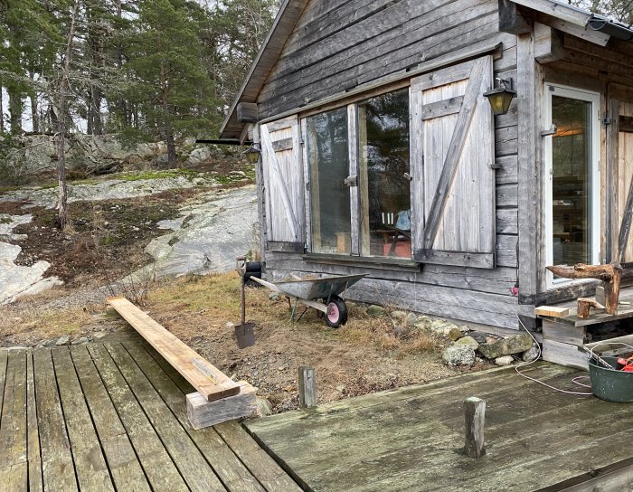 Grånat timmerhus vid havet med början av en altanbyggnation i förgrunden, tryckimpregnerade träplank och verktyg synliga.