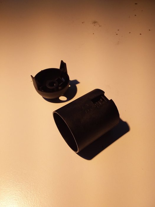 Två delar av en svart plastkomponent som verkar ha brutits isär, på en ojämn ljus yta.