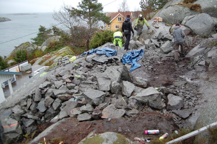 Arbetare spränger och hanterar sten manuellt på en bergig byggplats med havsutsikt.