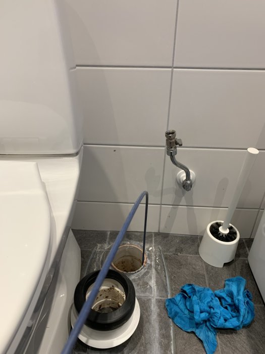 Toalettstol med öppen avloppsrörkoppling på badrumsgolv, blå trasa och rengöringsutrustning.