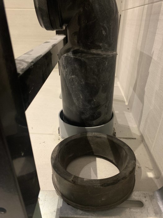 Cisternanslutning med svart böjt avloppsrör och en lös gumminippel på ett vitt kaklat golv.
