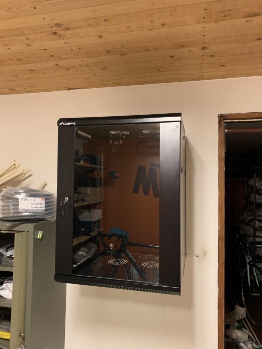 Nätverksskåp installerat i öppning i vägg med synliga kablar och en cykel reflekterad i glasdörren.