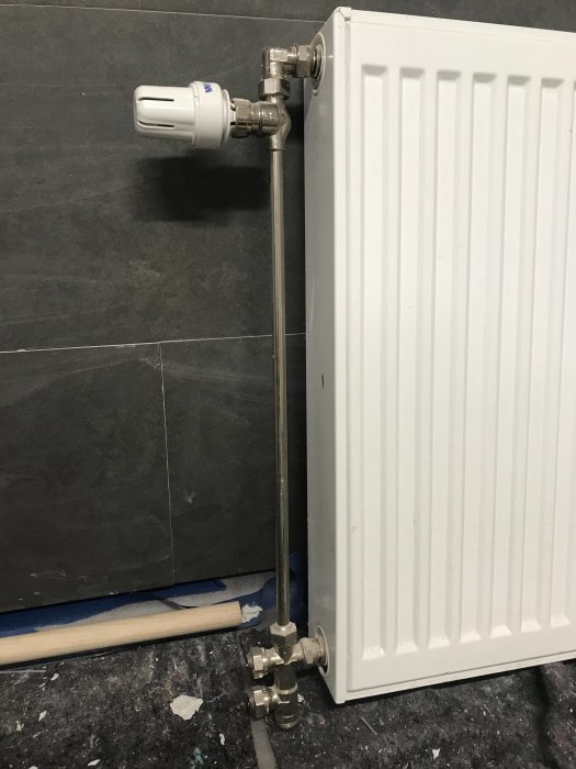 Vit radiator 60x60 cm med termostat och röranslutningar mot mörkgrå vägg och svart golv.