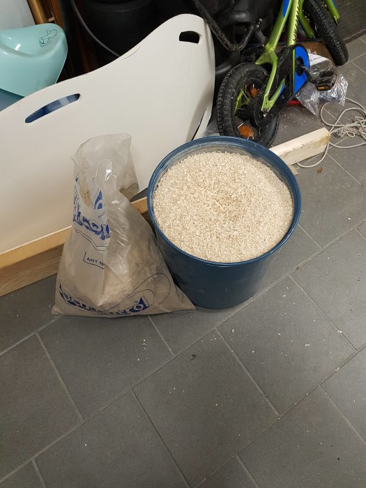 Blå spåntunna fylld med träspån bredvid en vit sopsäck i ett rörigt garage.
