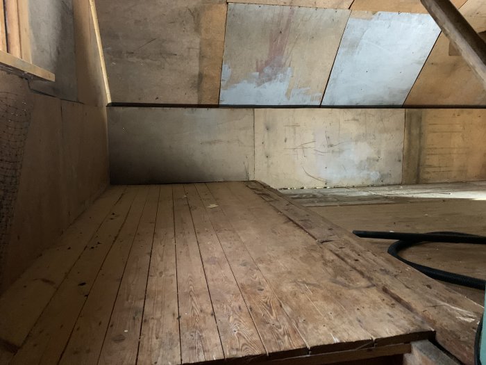 Trägolv och väggar i ett tomt förrum med synliga elledningar och dammsugarslang på golvet.