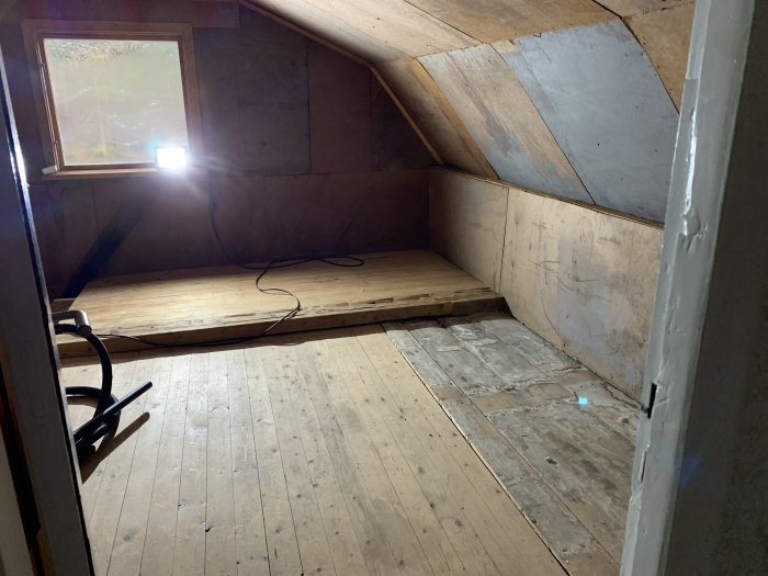Delvis renoverat rum med trägolv och plywoodväggar, öppen dörr och verktyg på golvet.