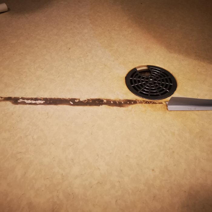 Spår efter borttagen T-list på badrumsgolv med gulaktig matta och en svart golvbrunn, bredvid en spackelspade.