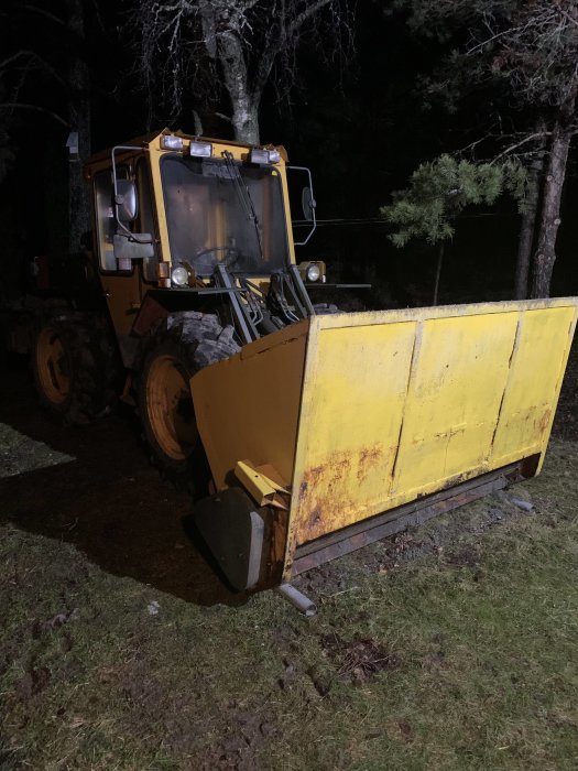 Gul vägmaskin med skopa parkerad utomhus på kvällen.