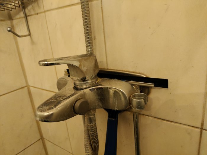 Blandare med duschslang i en kaklad dusch, med tecken på kalkavlagringar och bruksslitage.