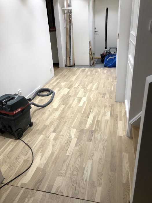 Nyinstallerat ljus parkettgolv i en hall med en dammsugare och byggmaterial i bakgrunden.
