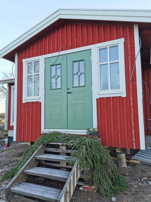 Grön ytterdörr med kod S5020-G30Y på röd träfasad, omgiven av vit fönsterkarm och trätrappa.
