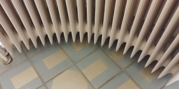 Gammal, smutsig radiator ovanpå ett fläckigt linoleumgolv med mönster i beige och blått.