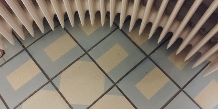 Gammaldags radiator ovanför ett mönstrat kakelgolv i beige och grått.