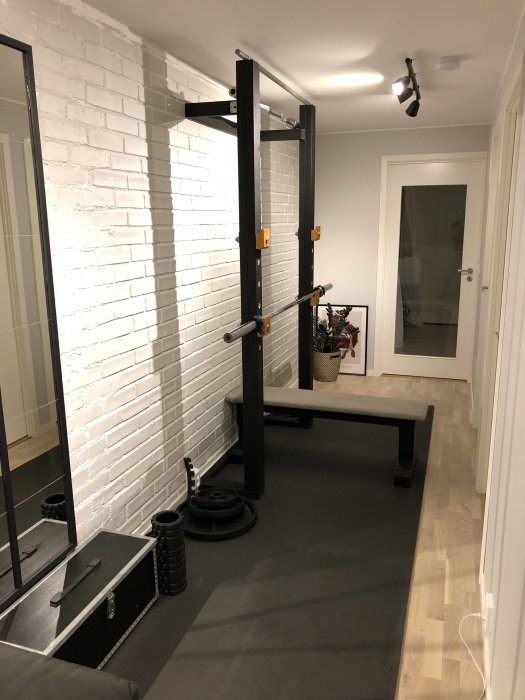 Hemmagym med squat rack, träningsbänk och vikter i en inomhuskorridor med vit tegelvägg.