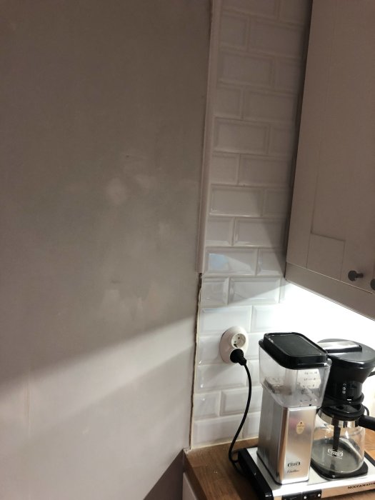 Vägg i kök med ojämn avslutning vid kakel, synliga spackelspår och eluttag, med en kaffebryggare i förgrunden.