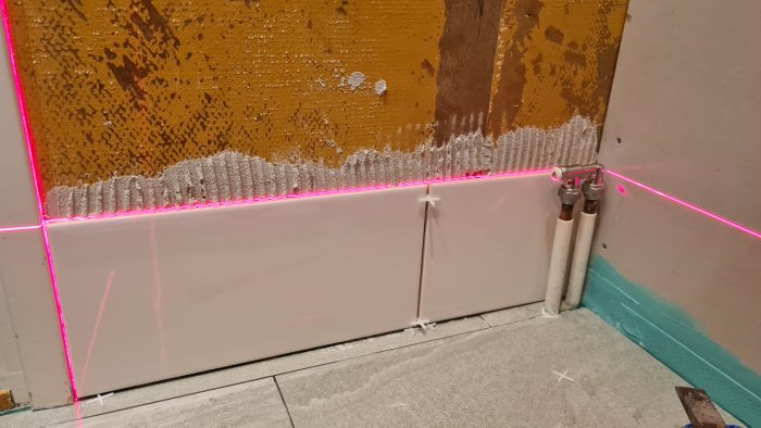 Kakelsättning med rosa laserlinje för nivåkontroll och första raden av vita kakelplattor installerade ovanför grå fix på vägg.