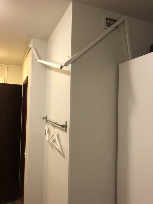 Felaktigt monterad klädhängare hänger lös från en vit vägg vid sidan av en stängd dörr.