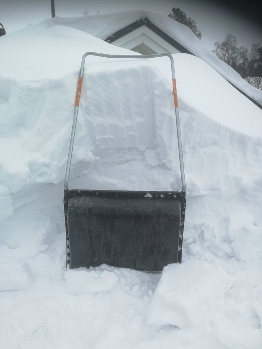 Snöskottning framför garage med tjocka snödrivor och ett snötäckt sadeltak i bakgrunden.