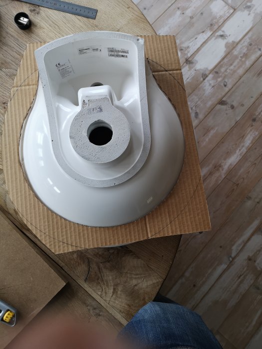 Kartongmall placerad ovanpå en vit toalettstol för att kontrollera passformen, med rullmått och trägolv i bakgrunden.