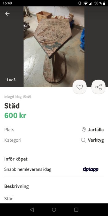 Använd, rostig städ i metall på en verkstads golv, sedd i profil med andra arbetsredskap i bakgrunden.